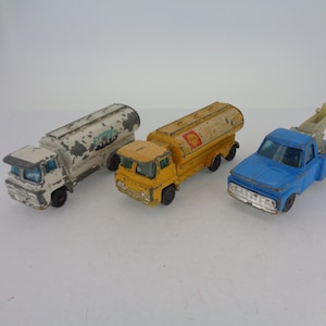 May include: Three vintage toy trucks, a white tanker truck, a yellow tanker truck, and a blue tow truck. The trucks are all in good condition, with some wear and tear from age.