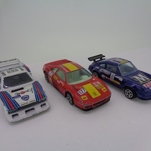 May include: Three toy race cars, a white and blue Lancia, a red Ferrari, and a blue Porsche. The cars are all in scale model form and feature sponsor logos and racing numbers.