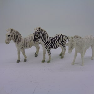 May include: Three plastic zebra figurines, one with black and white stripes, one with white and gray stripes, and one with white and gray stripes. The figurines are standing on a white surface.