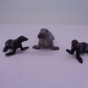 May include: Three plastic animal figurines: a gray walrus with ivory tusks, and two black sea lions. The walrus is facing forward, while the sea lions are facing to the left and right.