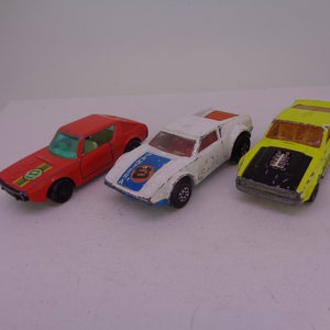May include: Three toy cars, a red car with a green and white stripe, a white car with a blue stripe and red accents, and a yellow car with a black hood and engine.