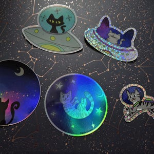 May include: A collection of holographic stickers featuring cats in space-themed designs. The stickers include cats in UFOs, against starry backgrounds, and in astronaut suits. The color palette includes blues, greens, and purples, with a black background.