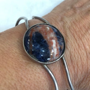 May include: A silver cuff bracelet with a large, oval stone. The stone displays a mix of blue, brown, and white hues. The bracelet has a simple design, with the stone set in a silver bezel and attached to the cuff, suitable for a wrist.