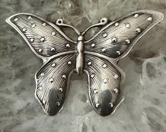 May include: A silver-colored butterfly-shaped brooch. The brooch has detailed wing patterns with small, raised dots. The butterfly's body is slender, and the wings are spread wide. The brooch is likely made of metal and is set against a light-colored background.