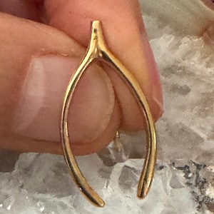 May include: A gold-toned wishbone pendant with a smooth, polished metallic finish. The wishbone features a classic bifurcated shape with two curved arms meeting at a single point at the top, creating a symmetrical and slender silhouette.