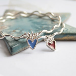 May include: Two silver bangle bracelets with a wavy design. One bracelet has a blue enamel heart charm and the other has a red enamel heart charm.