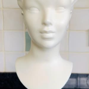 May include: A white, vintage-style mannequin head with closed eyes and styled hair. The bust is mounted on a black stand atop a white, circular base with a red light, likely for display or photography.