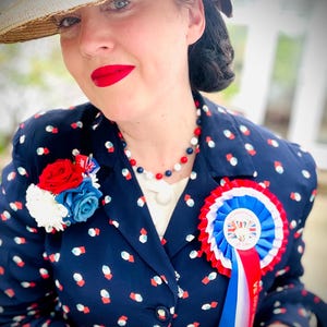 Könnte beinhalten: Eine Frau trägt eine marineblaue Jacke mit einem rot-weiß-blauen Muster. Sie trägt darunter ein weißes Hemd und ein rot-weiß-blaues Band mit einer Rosette. Die Rosette hat eine Union Jack und den Text "Victory Day" darauf. Sie trägt auch einen weißen Hut mit einem roten Band.