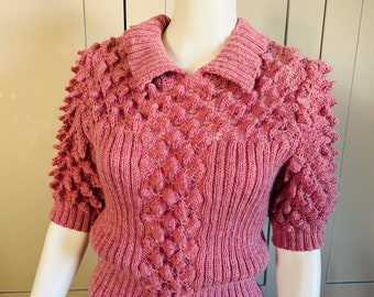 May include: A pink knitted sweater with a textured design and a collar. The sweater has short sleeves and a fitted silhouette.