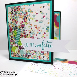 May include: A handmade greeting card with a confetti theme. The card features a black border, a teal inner border, and a white background filled with colorful confetti. The phrase "Cue the confetti" is written on a white banner, and party horn illustrations are included.