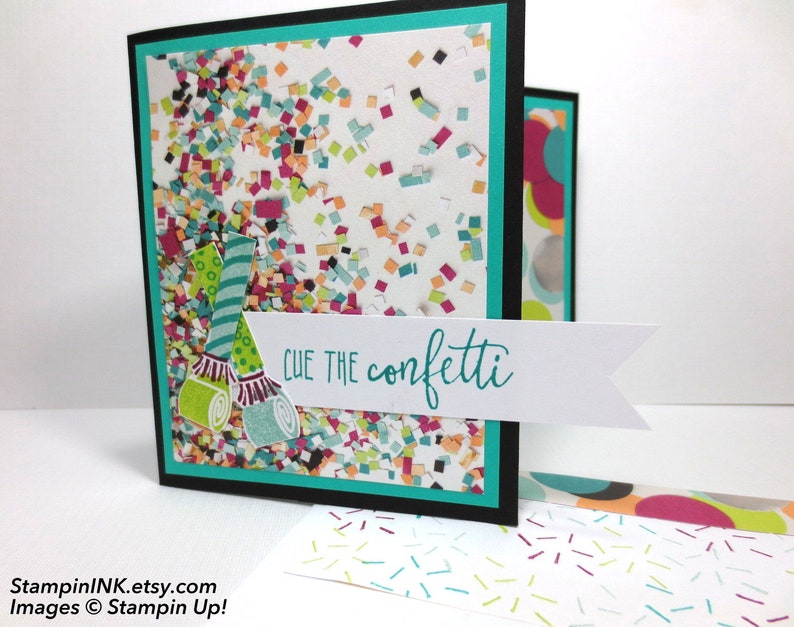 May include: A handmade greeting card with a confetti theme. The card features a black border, a teal inner border, and a white background filled with colorful confetti. The phrase "Cue the confetti" is written on a white banner, and party horn illustrations are included.