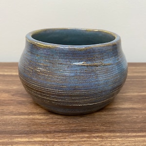 May include: A handmade ceramic bowl with a rounded shape and a blue glaze. The bowl has horizontal lines etched into the surface and a gold rim. The bowl is sitting on a wooden surface.