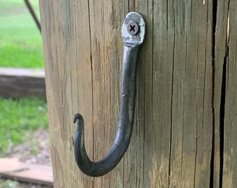 May include: A black metal hook is screwed into a weathered wooden post. The hook is bent into a J shape and has a small screw head at the top.