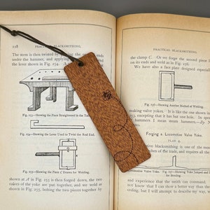 Könnte beinhalten: Nahaufnahme eines Holz-Lesezeichens mit einem geschnitzten Design. Das Lesezeichen ist braun mit einer hellbraunen Maserung. Das Design ist ein stilisiertes Blumenmuster mit einer Biene in der Mitte. Das Lesezeichen liegt auf einem geöffneten Buch mit Text über Schmiedekunst.