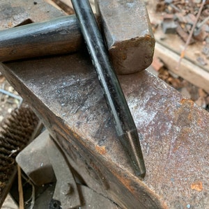 May include: A black metal punch tool with a pointed tip, resting on a metal anvil next to a black-handled sledgehammer.