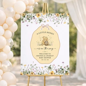 May include: A rectangular baby shower welcome sign on a gold easel, featuring a central yellow scalloped label with a bear illustration, honey pot, and the text A LITTLE HONEY IS ON THE WAY, AMANDA & CHARLES, 10-12-26, surrounded by floral borders.