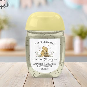 May include: A clear hand sanitizer bottle with a pale yellow cap and a white label featuring a cartoon bear with a honey pot, the text A LITTLE HONEY is on the way, AMANDA & CHARLES' BABY SHOWER, and the date 06.12.27 printed on the front.