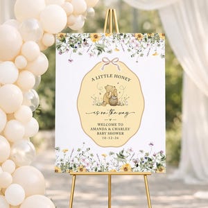 May include: A rectangular baby shower welcome sign on a gold easel, featuring a central yellow scalloped label with a bear illustration, honey pot, and the text A LITTLE HONEY IS ON THE WAY, AMANDA & CHARLES, 10-12-26, surrounded by floral borders.