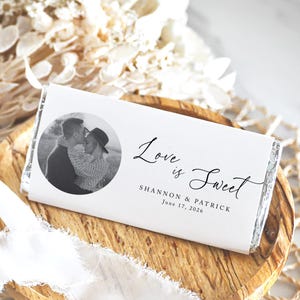 May include: A white chocolate bar wrapper with the text "Love is Sweet" and a black and white photo of a couple. The names Shannon & Patrick and the date June 17, 2026 are also printed on the wrapper. The wrapper is on a wooden plate.