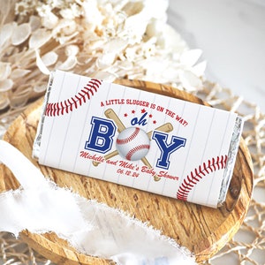May include: A white chocolate bar wrapper with a baseball theme. It says "A little slugger is on the way! Oh Boy!" with a baseball and bats. It also says "Michelle and Mike's Baby Shower 06.12.24".