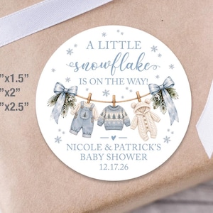 May include: A round sticker featuring a blue and white winter theme with the text A LITTLE snowflake IS ON THE WAY!, NICOLE & PATRICK'S BABY SHOWER, and 12.17.26. It shows baby clothes on a clothesline, available in 1.5x1.5, 2x2, and 2.5x2.5 inches.