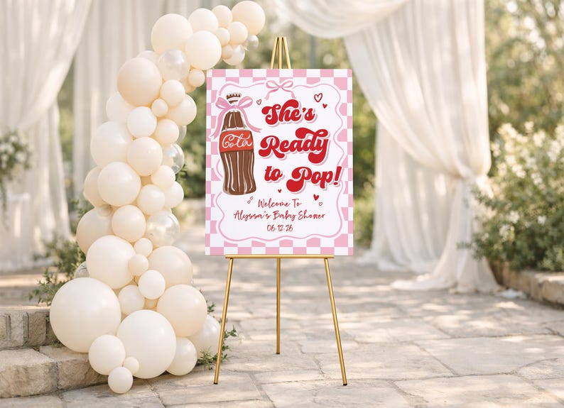 May include: A baby shower welcome sign on a gold easel. The sign is pink and white with a checkerboard pattern and features a brown soda bottle illustration and the text "She's Ready to Pop!" with the baby shower date.