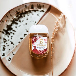May include: A small, clear hand sanitizer bottle with a white cap, decorated with a label featuring a red dress, flowers, butterflies, and the text "15 Christina Mi Quincea&ntilde;era 10.12.25". The bottle sits on a brown and white speckled plate.