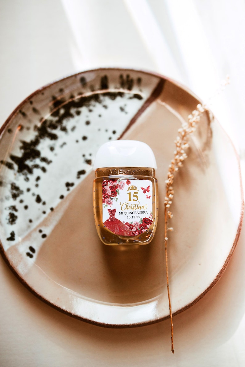 May include: A small, clear hand sanitizer bottle with a white cap, decorated with a label featuring a red dress, flowers, butterflies, and the text "15 Christina Mi Quincea&ntilde;era 10.12.25". The bottle sits on a brown and white speckled plate.