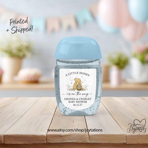 May include: A clear plastic hand sanitizer bottle with a light blue cap and a white label featuring a cartoon bear holding a honey pot, the text A LITTLE HONEY is on the way, AMANDA & CHARLES' BABY SHOWER, and the date 06.12.27.
