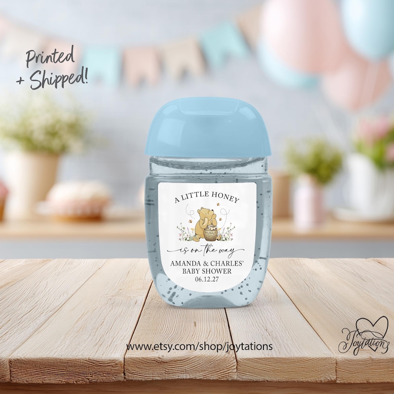 May include: A clear plastic hand sanitizer bottle with a light blue cap and a white label featuring a cartoon bear holding a honey pot, the text A LITTLE HONEY is on the way, AMANDA & CHARLES' BABY SHOWER, and the date 06.12.27.
