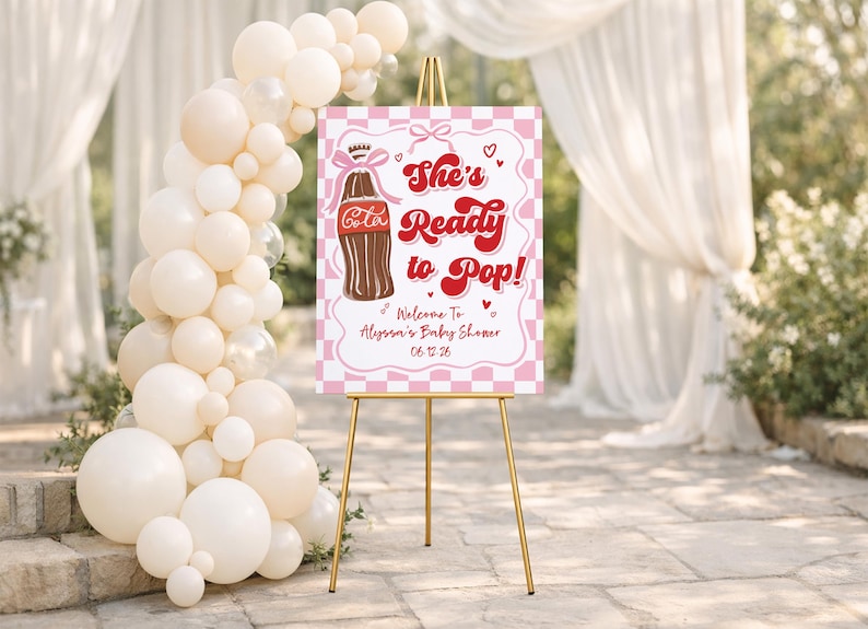 May include: A baby shower sign on a gold easel. The sign is pink and white with the text "She's Ready to Pop!" and a cartoon cola bottle. A balloon arch and draped fabric are in the background.