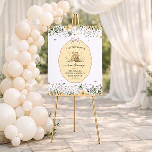 May include: A rectangular baby shower welcome sign on a gold easel, featuring a central yellow scalloped label with a bear illustration, honey pot, and the text A LITTLE HONEY IS ON THE WAY, AMANDA & CHARLES, 10-12-26, surrounded by floral borders.