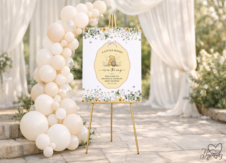 May include: A rectangular baby shower welcome sign on a gold easel, featuring a central yellow scalloped label with a bear illustration, honey pot, and the text A LITTLE HONEY IS ON THE WAY, AMANDA & CHARLES, 10-12-26, surrounded by floral borders.
