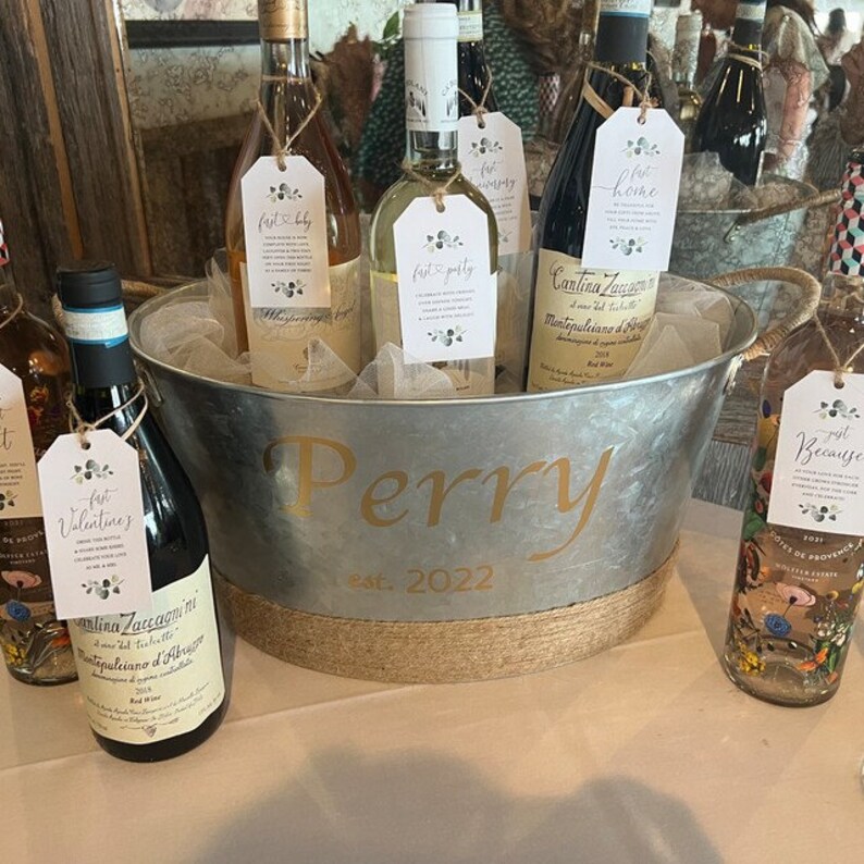 May include: A silver metal ice bucket with a brown rope trim, personalized with the name "Perry" and the date "Est. 2022". The bucket is filled with various bottles of wine, each with a personalized tag attached.