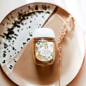 May include: A small, clear hand sanitizer bottle with a white cap, featuring a gold dress design and the text "15 Christina Mi Quinceanera 10.12.25." The bottle sits on a decorative plate with brown and white accents, with a dried flower stem.