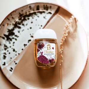May include: A small, clear hand sanitizer bottle with a white cap, featuring a label with a burgundy quincea&ntilde;era dress design and the name "Christina". The bottle sits on a speckled ceramic plate.