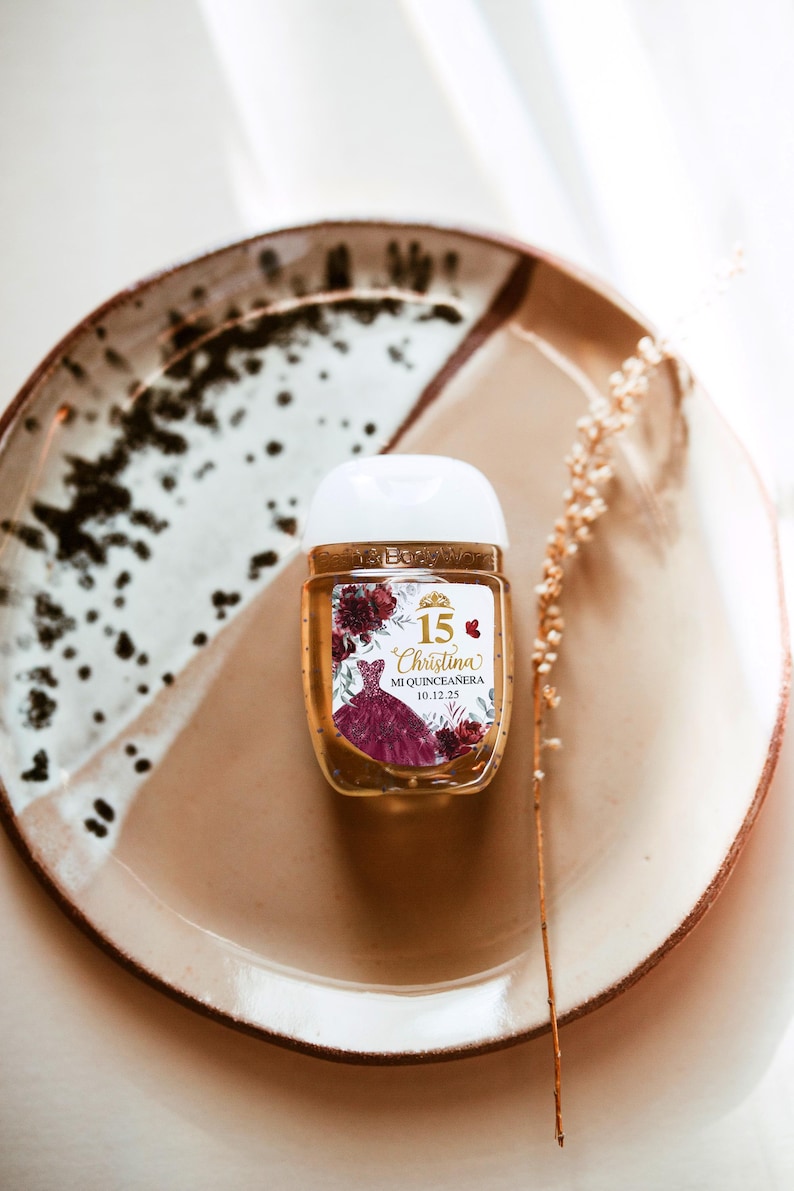 May include: A small, clear hand sanitizer bottle with a white cap, featuring a label with a burgundy quincea&ntilde;era dress design and the name "Christina". The bottle sits on a speckled ceramic plate.