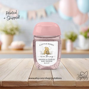 May include: A clear plastic hand sanitizer bottle with a light pink cap and a custom label featuring a Winnie the Pooh illustration, the text A LITTLE HONEY is on the way, AMANDA & CHARLES' BABY SHOWER, and the date 06.12.27 on a wooden surface.