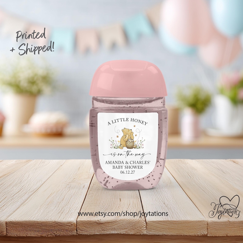 May include: A clear plastic hand sanitizer bottle with a light pink cap and a custom label featuring a Winnie the Pooh illustration, the text A LITTLE HONEY is on the way, AMANDA & CHARLES' BABY SHOWER, and the date 06.12.27 on a wooden surface.