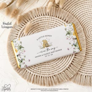 May include: A rectangular chocolate bar wrapper featuring a white background with a floral border and a central illustration of Winnie the Pooh holding a honey pot. The text reads A LITTLE HONEY is on the way AMANDA & CHARLES' BABY SHOWER 10-12-26.