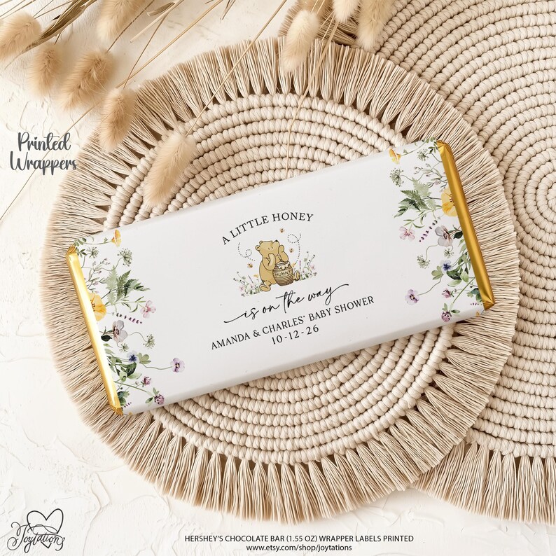 May include: A rectangular chocolate bar wrapper featuring a white background with a floral border and a central illustration of Winnie the Pooh holding a honey pot. The text reads A LITTLE HONEY is on the way AMANDA & CHARLES' BABY SHOWER 10-12-26.
