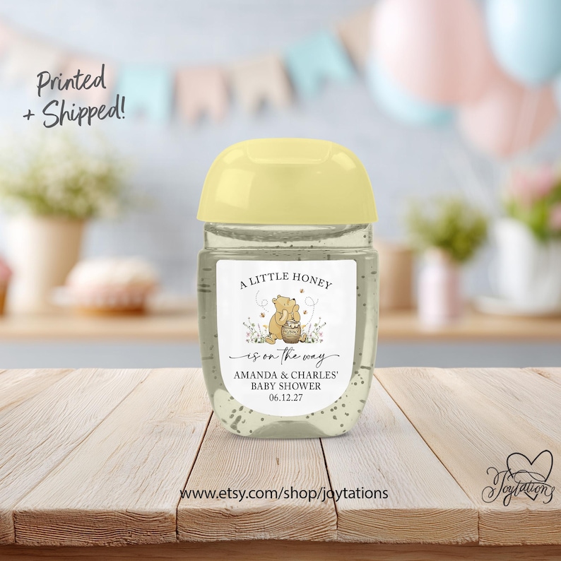 May include: A clear hand sanitizer bottle with a pale yellow cap and a white label featuring a cartoon bear with a honey pot, the text A LITTLE HONEY is on the way, AMANDA & CHARLES' BABY SHOWER, and the date 06.12.27 printed on the front.