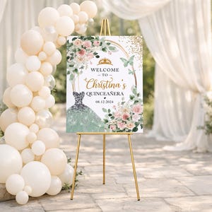 May include: A welcome sign for Christina's Quincea&ntilde;era, featuring a gold easel. The sign displays text and a dress illustration, surrounded by floral and butterfly accents. A balloon arch and draped fabric create a festive backdrop.