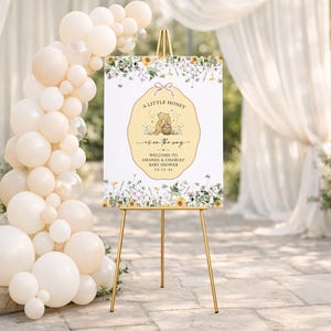May include: A rectangular baby shower welcome sign on a gold easel, featuring a central yellow scalloped label with a bear illustration, honey pot, and the text A LITTLE HONEY IS ON THE WAY, AMANDA & CHARLES, 10-12-26, surrounded by floral borders.
