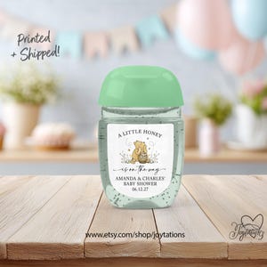 May include: A clear plastic hand sanitizer bottle with a light green flip-top cap. The label features a Winnie the Pooh illustration, the text A LITTLE HONEY is on the way, AMANDA & CHARLES' BABY SHOWER, and the date 06.12.27.