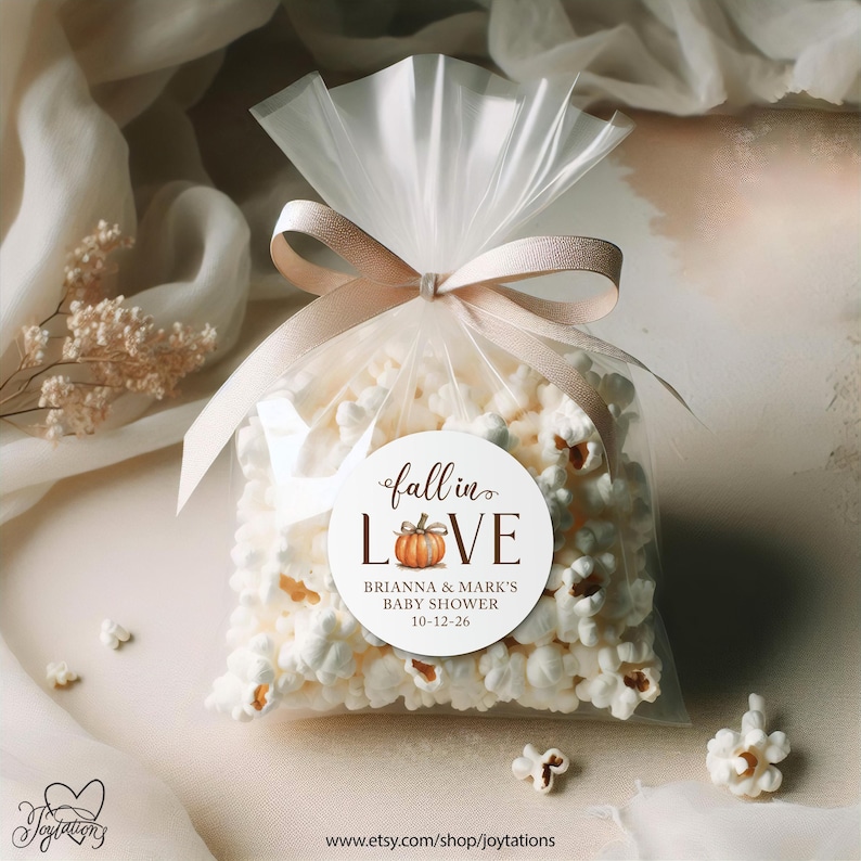 May include: A clear plastic bag filled with white popcorn, tied with a light beige ribbon. A circular white label on the front features the text "fall in LOVE" with a small pumpkin illustration, "BRIANNA & MARK'S BABY SHOWER", and "10-12-26".