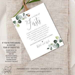 May include: A white tag with green eucalyptus leaves and the text "A Year of Firsts" with a basket of wine for a special pair. The text also includes "Congratulations Angela & Jamie!" and "Rachel and Kayla".