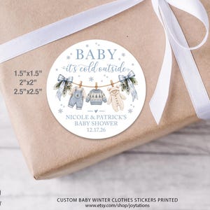 May include: A round white sticker featuring the text BABY it's cold outside, Nicole & Patrick's Baby Shower, and 12.17.26. The design shows baby clothes on a clothesline with blue bows, snowflakes, and available sizes 1.5x1.5, 2x2, and 2.5x2.5 inches.