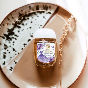 May include: A small, clear hand sanitizer bottle with a white cap, featuring a label with the text "15 Christina Mi Quincea&ntilde;era 10.12.25". The label includes a purple dress illustration and floral accents. The bottle sits on a brown and white plate.
