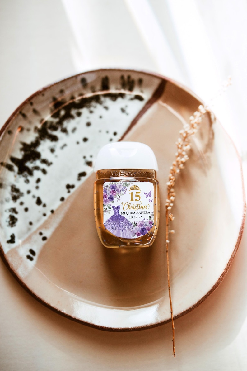 May include: A small, clear hand sanitizer bottle with a white cap, featuring a label with the text "15 Christina Mi Quincea&ntilde;era 10.12.25". The label includes a purple dress illustration and floral accents. The bottle sits on a brown and white plate.